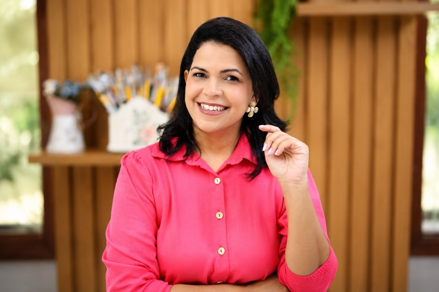 Ana Laura Rodrigues sorrindo, de camisa rosa, com seus pincéis de pintura ao fundo